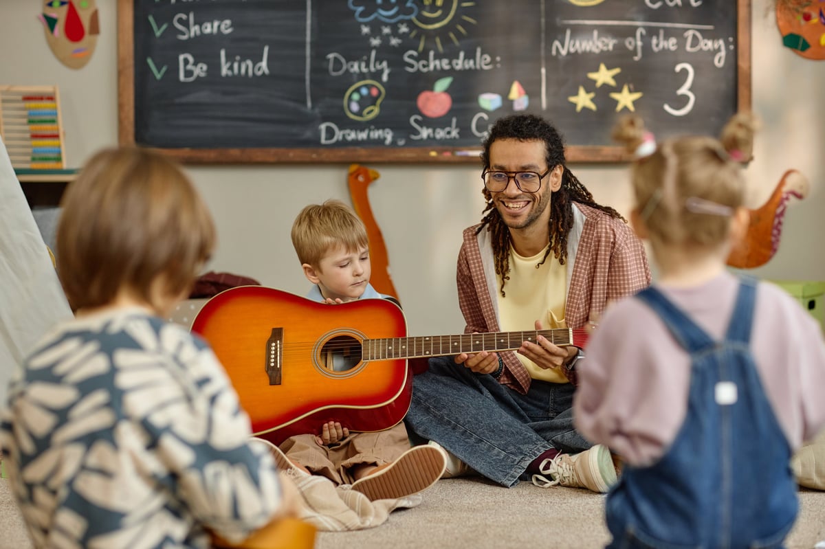 Music teacher sitting with a group of children presenting an acoustic guitar, children listening and watching during a music lesson